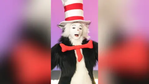 A man in a furry cat suit. He is wearing red and white striped hat and a red neck tie..