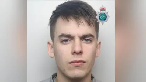 A mugshot of a man with short brown hair and wearing a grey top.