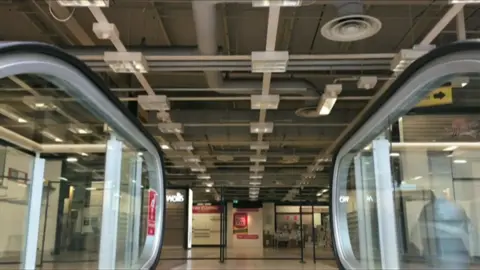 Archive shot showing the interior of the Debenhams store. The image shows the top of an escalator with large glass panels in the background. There are posters on the wall.