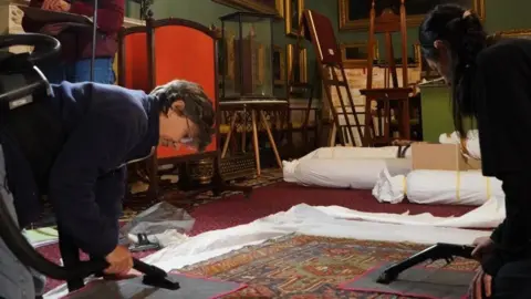 National Trust/ Lucy Fenna Two people in the room of a stately home surrounded by antique furniture bent over an antique rug with special mats and small hoovers.