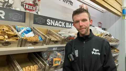 Jim Connolly/BBC Matthew at Paw Prints. He has brown hair and a short beard, wearing a black jumper with the company name on it in white. He stands in front of a stand of dog treats.