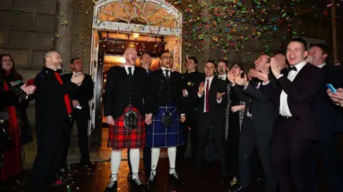 Getty Images Two men in kilts getting married. A crowd is cheering and laughing, clapping their hands as confetti falls over them all.