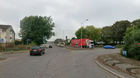 Google Street view of a roundabout and a straight road. It is a cloudy day and there are cars traveling on the road.