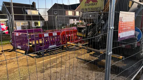 John Devine/BBC Work taking place on the site in Broadway. There are various fences positioned around a hole, with a lorry parked on site.