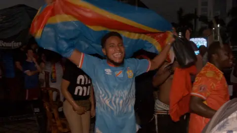 A fan celebrates. He is wearing a blue football shirt and holding up the Congolese flag behind his head