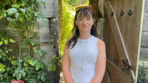 A woman in a white vest and long dark hair stands beside an old stone doorway with a wooden door. She has white headphones hanging around her neck and sunglasses perched on her head. Plants grow up the side of the stone wall. Behind her and through the door leads to a very sunny garden.