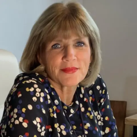 Dr Tricia Shaw A woman is smiling lightly at the camera for a head and shoulders shot. She has shoulder length dark blonde hair with a full fringe and is wearing red lipstick and a navy jumper with white, red, yellow and blue dots on it. She is sitting on a beige chair.