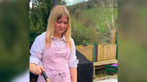 A teenage girl with straight blonde hair past her shoulders cooks food on a BBQ in a garden. She is wearing a white, rolled up shirt and a pink apron. She is smiling slightly.