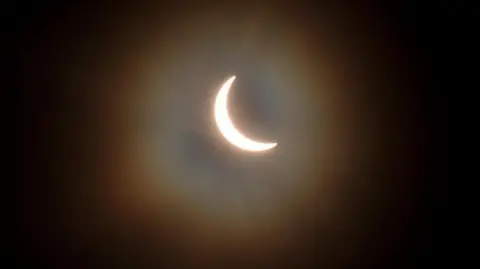 Getty Images The sun is pictured during a partial solar eclipse. The sky is dark and cloudy. The sun is a thin arc in the sky.