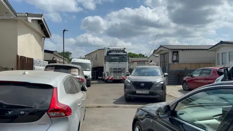 A mobile home sits on the back of a large lorry and is transported through the Traveller site