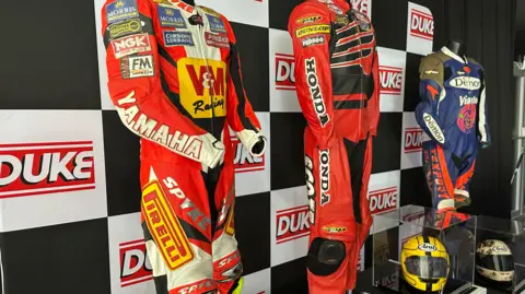 Three sets of colourful leathers feature on mannequins. In front there are helmets in clear boxes. They stand in front of a black and white chequered backdrop, which also features a red and white Duke logo.