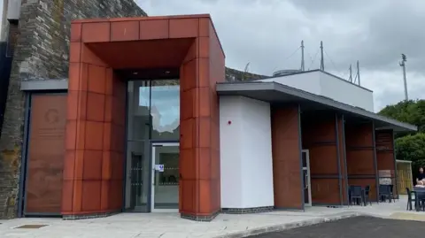 The outside of the museum is made of a red-brown material. It's built close to a large grey stone wall. 