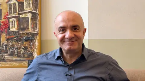 Imad Alarnab, who is bald and wearing a blue shirt, looking at the camera and smiling. He is sat on a sofa in front of a wall, with a painting on the left behind him.