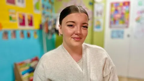 Martin Giles/BBC A woman is sitting in a room for children. It has bright coloured walls painted blue, yellow and green. They have pictures, photos and children's wall charts on them. Isabella is wearing a white top and matching cardigan. She is smiling at the camera and has her hair tied back.