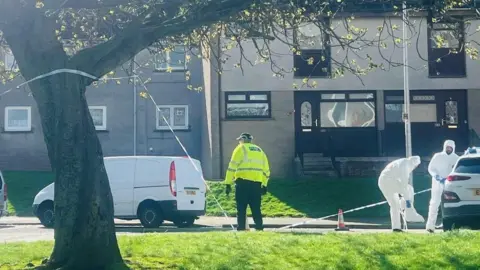 Two people in white forensic suits and blue gloves, outside a building, with blue and white police tape in the foreground, and a police officer in a yellow jacket.