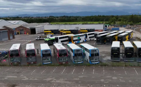 PA Media A large car park outside a warehouse with at least a dozen buses in various colours parked in chevron formations
