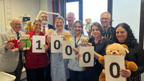 Staff stand in a hospital ward holding teddy bears and sheets of paper displaying the number 10,000