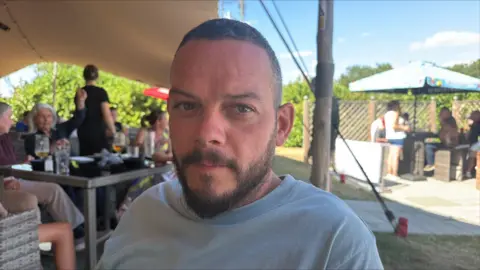 Richard Knights/BBC A man is sitting in a pub garden, which is packed with people. He is wearing a light grey T-shirt, has a short beard and is looking directly at the camera.