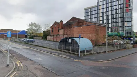 Google Street view image of the proposed plot. A car park by a junction with red brick building in the background and a round shelter to the front