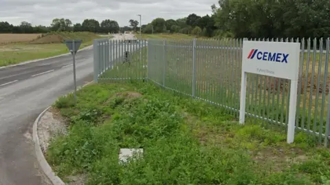 An road entrance to a concrete plant. The road runs through the middle and a silver metal fence is along the perimeter. The gates are open and a patch of vegetation is in front of the fence with a white sign that reads "Cemex". 