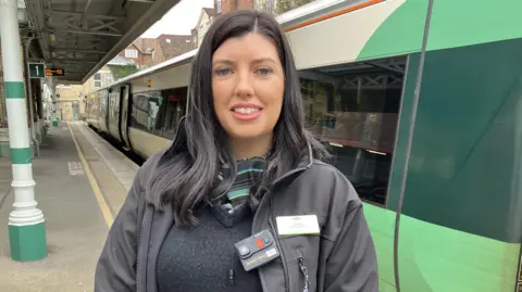Leanne Rinne/BBC A woman with long black hair standing in front of a green train on a platform. She is smiling and wearing Southern Railway uniform. 