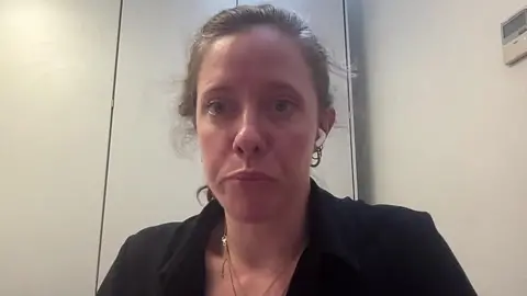 A woman in her 30s with light brown hair tied back and a black jacket speaks into a camera