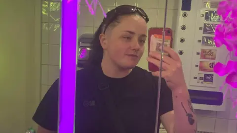 Amber Stebbings wearing a dark T-shirt, taking a photo of herself in the mirror with a red phone. She has tattoos on the arm she is holding the phone up with. In the background is a wall-mounted vending machine. 