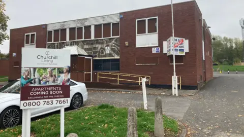LDRS A 1970s flat-roofed brick building with boarded-up windows and a "sold" sign on the wall. On the grass verge, there is a sign advertising the new Churchill Living apartments "coming soon".