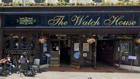 Google Picture of The Watch House pub with signage written in gold on a blue frontage. Outside the pub there are hanging baskets and a few people sat the left with drinks. 