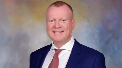 Professional studio portrait of Dennis Bacon wearing a navy suit, white shirt and muted red tie, facing the camera against a softly blended blue, grey and beige background. The image is sharply focused with even lighting and a formal corporate style.