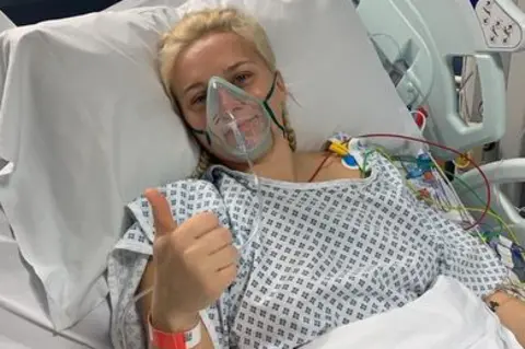 Courtney Evans in a hospital bed wearing an oxygen mask. She has blonde hair and is holding her thumb up to the camera.