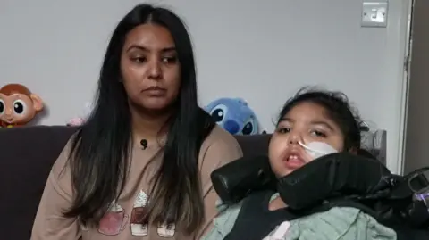 BBC A picture of mum Namla (left) with dark brown hair and daughter Sara (right) sitting in a wheelchair with a feeding tube in her nose.