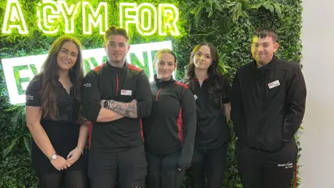 Everyone Active A group of young gym staff, all wearing black uniforms, pose for the camera inside Horfield Leisure Centre. Behind them is a wall of fake greenery with a neon sign in it