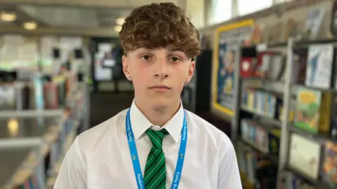 Sam Read/BBC Kohen looks at the camera without smiling. He has a short-sleeved white school shirt on and green/black striped tie, with a blue lanyard. He wears a gold stud earring in each ear.