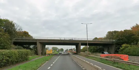 Google A Google street view image shows a bridge over a dual carriageway. A yellow road works sign and orange barriers can be seen on the carriageway.