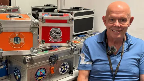 An older man with a bald head sits in a dressing room, surrounded by colourful orange, red, black and silver metal boxes decorated with Northern Soul club stickers. He wears a blue polo shirt and a black lanyard around his neck. A mirror is visible behind the boxes.