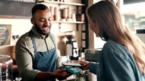 Getty Images A woman customer with long blonde hair and a denim shirt taps her phone onto a card machine which is being held by a worker in a white striped denim apron