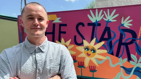 Chef Tony Church smiling for a picture with his arms folded in front of his chest in front of a colourful mural reading The Star. It is a sunny day.
