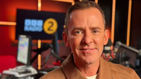 Scott Mills in a brown jacket in the radio studio