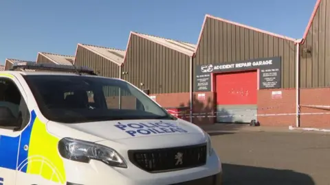 A police van sits outside a garage in East Kilbride, where a serious assault has taken place. Police tape is visible around the building.