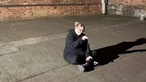 Michelle Liddle Michelle Liddle is holding a pendulum while trying to communicate with paranormal forces. She is wearing a black hoodie and say cross-legged on the floor.