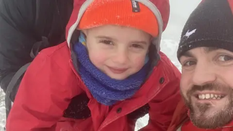A four-year-old boy wearing a red coat, blue snood, orange hat and black gloves, is pictured at the summit of a mountain alongside a man in an orange coat and black hat, with a short brown beard. The boy is being held up on to the trig pillar by a person in a black coat whose face can't be seen. Both the boy and the man are smiling. It is misty and snowy in the background and there is snow on the boy's gloves and hat.