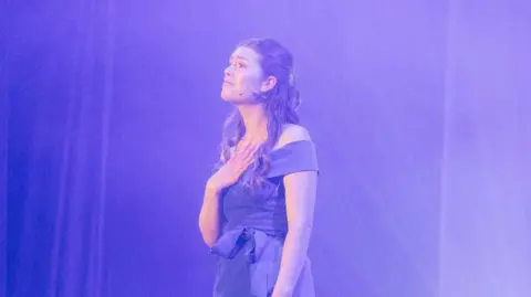An image of a young woman with long dark hair wearing a purple sleeveless dress. She is standing side-on to the camera, with her left hand hanging down by her side and her right hand rested palm-side down on her chest. She is wearing a small headset mic.
