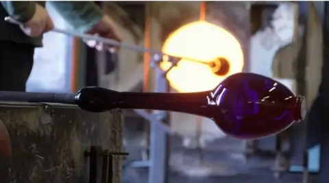 Photo inside Bristol's blue glass factory. This picture shows a blue vase being shaped on a stick in the factory. In the background you can see a man putting a a long stick into furnace.