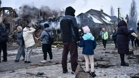 Getty Images A man holds a girl's hand and looks at buildings destroyed in a Russian drone and missile strike on Kyiv in February 2026
