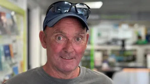 Richard stands in the pharmacy. He is wearing a grey T-shirt and a baseball cap with glasses balanced on top of his hat.