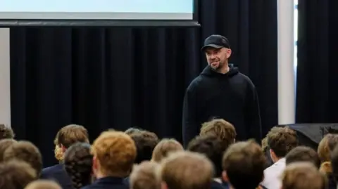 Seaford College A group of children had their heads and backs turned to the camera, and they are facing towards a stage and a projector screen. On the stage is Matt Pinkett, who wears a black hoodie and black baseball cap.