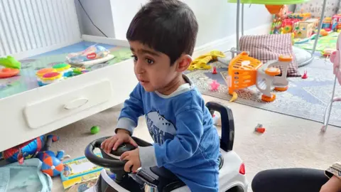 PA Media Eisa at home in a playroom, surrounded by toys is riding a play car. He is wearing a blue baby grow, has dark hair and eyes.