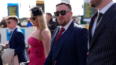 PA Media Darts star Luke Littler walks into the Cheltenham Festival, with his other guests either side. He is wearing a dark blue suit and waistcoat, a white shirt and dark pink tie and sunglasses