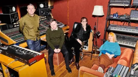 Tom Jackson Three men and a woman lounge in a recording studio. The woman has blonde hair and is sprawled out on an orange sofa. There are keyboards and mixing desks in the background